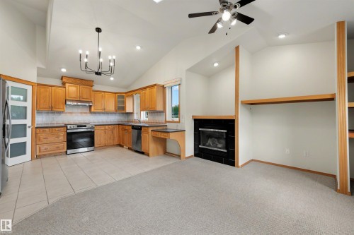 Open-concept living space with vaulted ceilings, a kitchen featuring wood cabinetry and stainless steel appliances, a black tile-surround fireplace, and recessed lighting - 15227 47 Street, Edmonton, AB 