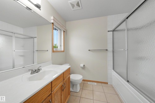 Bathroom featuring a wood-finish vanity with an integrated sink, a large wall-mounted mirror, and a shower-over-tub with sliding frosted glass doors - 15227 47 Street, Edmonton, AB 