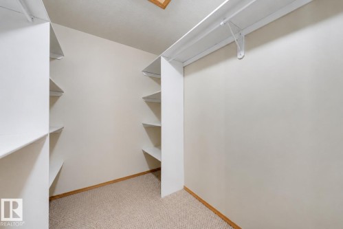 Walk-in closet featuring white built-in shelving, a garment rod, light beige wall paint, and carpet flooring with wood-finish trim - 15227 47 Street, Edmonton, AB 