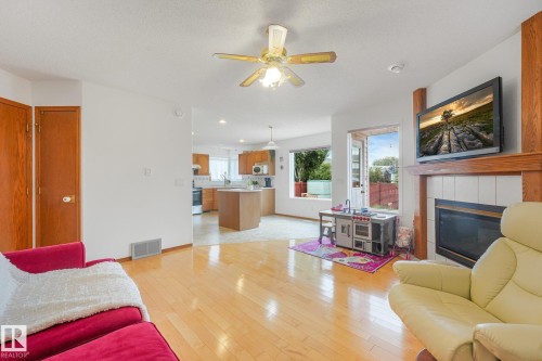 Hardwood flooring with a ceiling fan, a fireplace with a white tile surround and wood mantel, and a kitchen with wood-finish cabinetry - 1225 Kane Wynd Nw, Edmonton, AB - Indoor Photo Showing Living Room With Fireplace