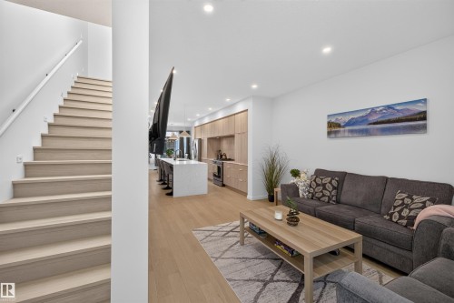 Open-concept living space with light wood-finish flooring, recessed lighting, a modern staircase with a white handrail, and a kitchen featuring light wood cabinetry and a white island with a built-in sink - 8551 81 Avenue, Edmonton, AB 