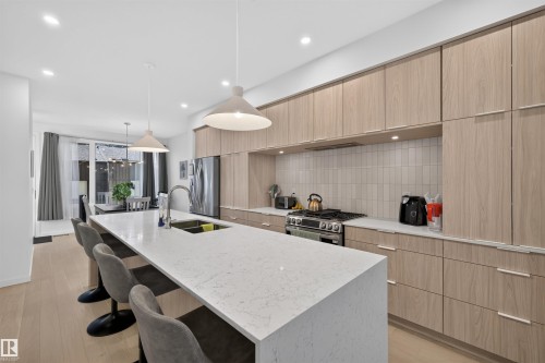 Modern kitchen featuring a large island with a light-toned countertop, light wood-finish cabinetry, stainless steel appliances, and wood-finish flooring - 8551 81 Avenue, Edmonton, AB 