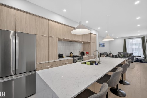 Open-concept kitchen featuring a large island with a light-toned countertop, stainless steel appliances, light wood-finish cabinetry, a gas range with a tiled backsplash, and recessed lighting - 8551 81 Avenue, Edmonton, AB 