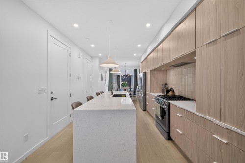 Open-concept kitchen featuring light wood-finish cabinetry, a large center island with a stone countertop, recessed lighting, stainless steel appliances, and wood-finish flooring - 8551 81 Avenue, Edmonton, AB 