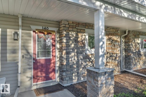 Entryway featuring a red paneled door with glass insert, stone veneer siding, and a covered porch with a stone-clad column - 140 655 Tamarack Road, Edmonton, AB 