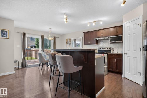 Open-concept kitchen and dining area featuring rich wood-finish flooring, dark wood cabinetry, stainless steel appliances, a white subway tile backsplash, and a breakfast bar with seating - 7858 170A Avenue, Edmonton, AB 