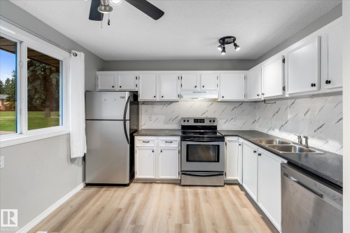 Light-filled kitchen featuring wood-finish flooring, white cabinetry with dark hardware, dark countertops, a marble-patterned backsplash, and stainless steel appliances - Fort Saskatchewan, AB 
