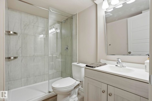 Full bathroom featuring a glass-enclosed shower with large format tile surround, a single vanity with an integrated sink, a rectangular framed mirror, and wood-finish flooring - 445 Watt Boulevard, Edmonton, AB 
