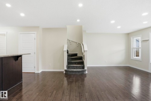 Spacious interior featuring dark wood-finish flooring, recessed ceiling lighting, a dark wood-finish island with a light-toned countertop, and a carpeted staircase with white railings - 445 Watt Boulevard, Edmonton, AB 