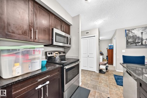 Kitchen featuring dark wood cabinetry, stainless steel appliances including a microwave and range, dark stone countertops, and square floor tiles - 417 1510 Watt Drive, Edmonton, AB 