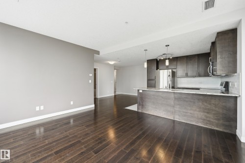 Open-concept living space featuring wood-finish flooring, a kitchen island with stone countertops, dark wood-finish cabinetry, stainless steel appliances, and a tiled backsplash - 528 5151 Windermere Boulevard, Edmonton, AB 