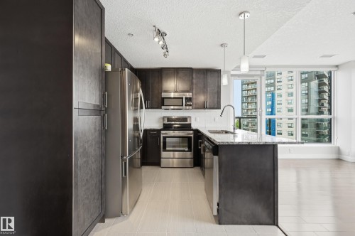 Kitchen featuring dark wood-finish cabinetry, stainless steel appliances, a center island with pendant lighting, and a window framing urban architecture - 528 5151 Windermere Boulevard, Edmonton, AB 