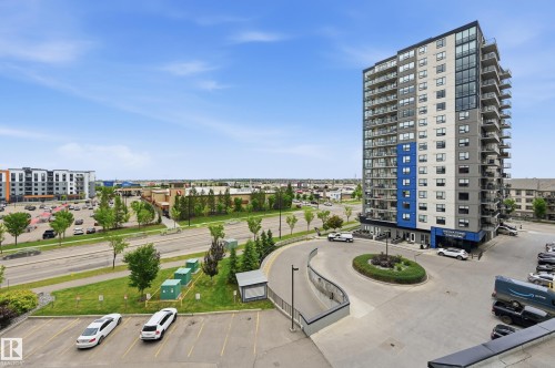 Modern high-rise building featuring a striking blue and gray exterior, multiple balconies, a circular driveway with landscaping, and an adjacent parking area - 528 5151 Windermere Boulevard, Edmonton, AB 