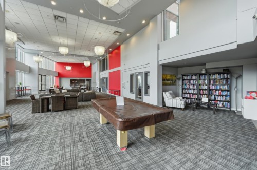 Spacious common area featuring patterned carpet flooring, a high ceiling with recessed lighting and pendant fixtures, an integrated lounge area, and a library section with built-in shelving - 528 5151 Windermere Boulevard, Edmonton, AB 