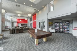 Spacious common area featuring patterned carpet flooring, a high ceiling with recessed lighting and pendant fixtures, an integrated lounge area, and a library section with built-in shelving - 
