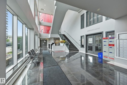Modern lobby featuring polished dark tile flooring, a reception desk, a multi-story atrium with a prominent staircase, full-height windows, and tenant mailboxes - 528 5151 Windermere Boulevard, Edmonton, AB 