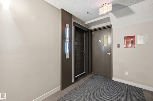 Hallway entry featuring a modern dark wood-finish door, light beige walls, and contemporary wall sconce lighting - 528 5151 Windermere Boulevard, Edmonton, AB 