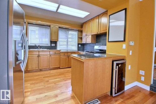 Kitchen featuring wood-finish flooring, natural wood cabinetry, a stainless steel refrigerator, a built-in beverage cooler, and a dark stone countertop - 11312 127 Street, Edmonton, AB 