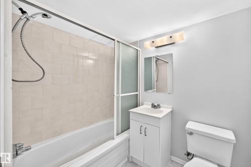 Bathroom featuring a white bathtub with tile surround, sliding frosted glass shower doors, a vanity with integrated sink, a wall-mounted mirror, and a modern light fixture - 11447 22 Avenue Nw, Edmonton, AB 