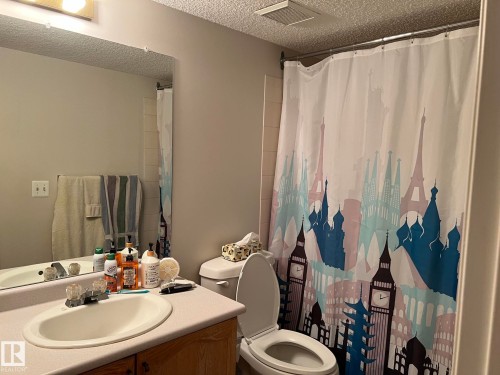 Bathroom featuring a single vanity with an oval sink, a wall-mounted mirror, and a built-in shower with a curtain rod - 207 18012 95 Avenue, Edmonton, AB - Indoor Photo Showing Bathroom