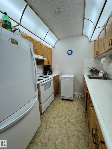Galley kitchen featuring light wood cabinetry with dark hardware, white countertops, a built-in double basin sink, white appliances, and patterned flooring - 207 18012 95 Avenue, Edmonton, AB 