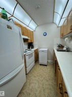 Galley kitchen featuring light wood cabinetry with dark hardware, white countertops, a built-in double basin sink, white appliances, and patterned flooring - 