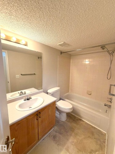 Bathroom featuring a vanity with wood-finish cabinetry and an integrated sink, a full-width mirror, a toilet, and a bathtub with tile surround - 207 18012 95 Avenue, Edmonton, AB 