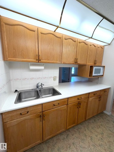 Kitchen featuring wood-finish cabinetry, a double basin stainless steel sink, a microwave alcove, white countertops, and a white tile backsplash - 207 18012 95 Avenue, Edmonton, AB 