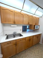 Kitchen featuring wood-finish cabinetry, a double basin stainless steel sink, a microwave alcove, white countertops, and a white tile backsplash - 