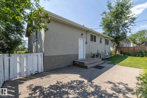 Rear exterior featuring a stucco finish, a concrete patio, a wooden deck with steps, a white entry door, and a grassed yard with mature trees - 5608 122 Avenue, Edmonton, AB 