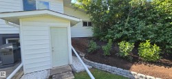 White vinyl siding exterior with a covered entry, established landscaping, and a tiered retaining wall - 