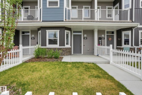 Townhome exterior featuring white picket fencing, an individual grassy area, a concrete walkway, and a covered porch entry - 36 150 Everitt Dr, St. Albert, AB - Outdoor With Balcony With Facade