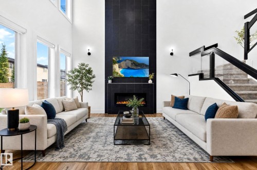 Living room featuring a two-story ceiling, large windows, wood-finish flooring, a contemporary fireplace with a dark tile surround, and a staircase with a glass and black metal railing - 423 42 Street, Edmonton, AB 
