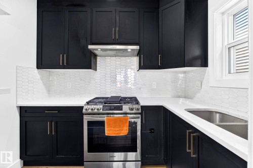 Contemporary kitchen featuring dark cabinetry with gold-tone hardware, a stainless steel range with overhead ventilation, white countertop surfaces, and a unique elongated hexagonal tile backsplash - 423 42 Street, Edmonton, AB 