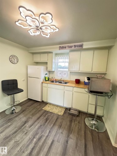 Kitchen featuring wood-finish flooring, white cabinetry, a double basin sink, and a floral-inspired ceiling light fixture - 4912 49 Avenue, Onoway, AB 