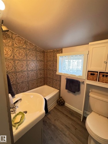 Full bathroom featuring a tub/shower combination with mosaic tile surround, a single vanity with an integrated sink, and wood-finish flooring - 4912 49 Avenue, Onoway, AB 