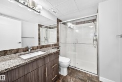Bathroom featuring a wood-finish vanity with an integrated rectangular sink and speckled countertop, a large wall-mounted mirror, a frameless glass shower enclosure, and dark stone-look floor tiles - 