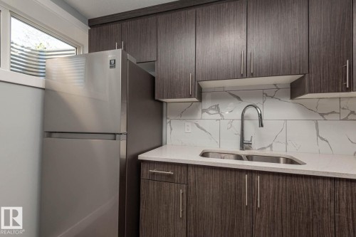 Contemporary kitchen featuring dark wood-finish cabinetry, a stainless steel refrigerator, white quartz-finish countertops, a dual basin stainless steel sink with a gooseneck faucet, and a large format tile backsplash with a grey vein pattern - 2635 Donaghey Crescent, Edmonton, AB 