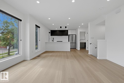 Bright open-concept living space with expansive windows, light wood-finish flooring, recessed lighting, and a modern kitchen featuring an island, dark cabinetry, and stainless steel appliances - 8903 94 Ave Nw, Edmonton, AB 