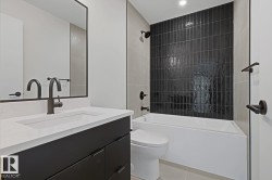 Contemporary bathroom featuring a dark tile shower surround, a white built-in tub, a dark vanity with a white countertop, and matte black fixtures - 