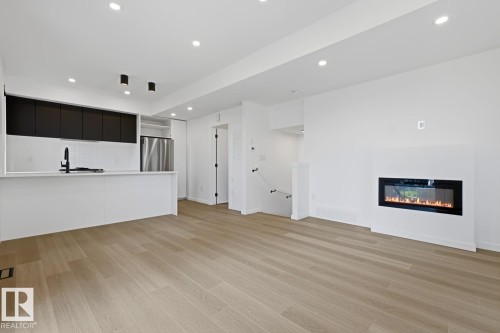 Open-concept living area featuring light wood-finish flooring, recessed ceiling lighting, a contemporary linear fireplace, and a kitchen island with a black matte faucet - 8903 94 Ave Nw, Edmonton, AB 