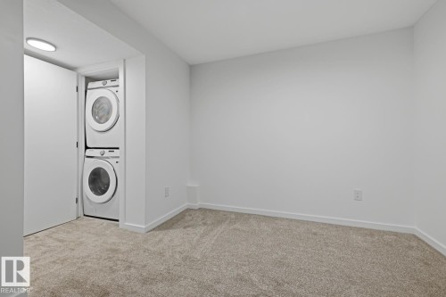 Dedicated laundry closet featuring a stacked washer and dryer unit, set within a neutral-toned room with carpet flooring and crisp white walls - 8903 94 Ave Nw, Edmonton, AB 