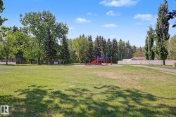 Expansive green space featuring a vibrant red and blue playground structure, mature trees, a paved walking path, and a white privacy fence bordering the perimeter - 