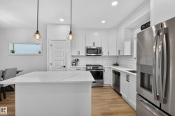 Modern kitchen featuring a large central island with a light-toned countertop, white cabinetry with matte black hardware, stainless steel appliances, and wood-finish flooring - 