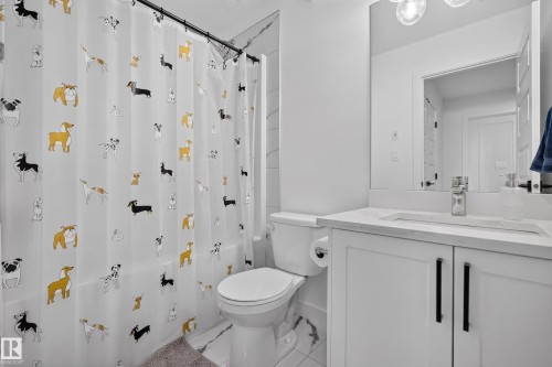 Bathroom featuring a white vanity with a recessed sink and quartz-style countertop, a wall-mounted mirror, a modern three-light fixture, and a toilet with a side-mounted roll holder - 548 Kleins Crescent, Leduc, AB 