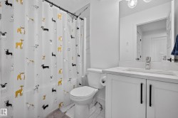 Bathroom featuring a white vanity with a recessed sink and quartz-style countertop, a wall-mounted mirror, a modern three-light fixture, and a toilet with a side-mounted roll holder - 