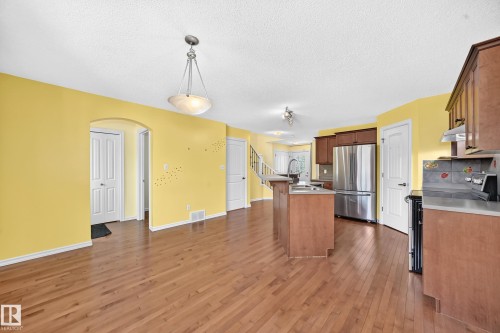 Open-concept kitchen and living area featuring wood-finish flooring, a central island with an integrated sink, stainless steel refrigerator, wood cabinetry, and an arched entryway - 211 59 Street, Edmonton, AB 