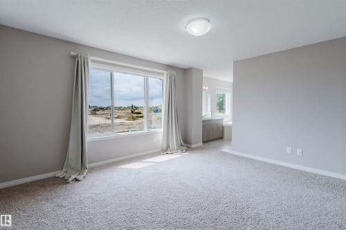Spacious carpeted room featuring a large window with white trim, neutral-toned walls, and a ceiling-mounted light fixture - 16627 18 Avenue, Edmonton, AB 