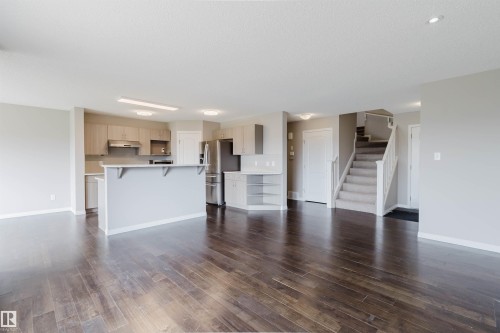 Open concept living area featuring dark wood-finish flooring, a kitchen with light wood cabinetry and stainless steel appliances, a kitchen island with a breakfast bar, and a carpeted staircase - 16627 18 Avenue, Edmonton, AB 