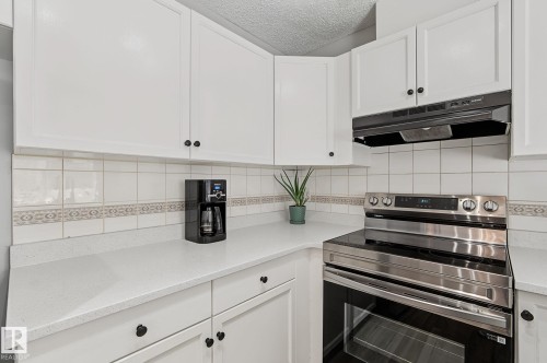 Kitchen featuring white cabinetry, solid surface countertops, white tile backsplash with decorative accent, and a stainless steel range with a built-in exhaust hood - 5 14603 Miller Boulevard, Edmonton, AB 